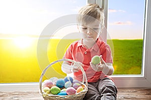 Positive kid holding basket full of easter eggs after egg hunt enjoying and sitting on the windowsill against background of a