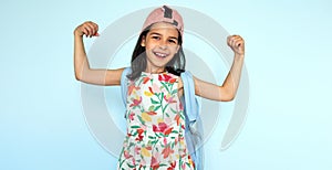 Positive kid smiling in dress, pink cap, and blue backpack looking at the camera showing her muscles against blue background.