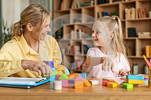 Positive kids psychology. Professional female therapist with notepad watching little girl playing with colorful blocks