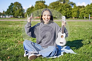 Positive korean girl sits in park, shows ukulele and rock on gesture, learns how to play musical instrument outdoors