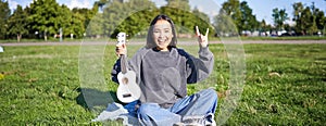 Positive korean girl sits in park, shows ukulele and rock on gesture, learns how to play musical instrument outdoors