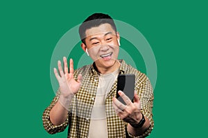 Positive korean man making video call via cellphone, wearing earphones and waving hello at webcamera, green background
