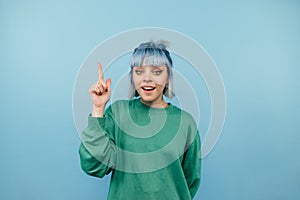 Positive lady with blue hair stands on a blue background with a happy face looking at the camera and showing finger up