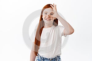 Positive little girl, child with long red hair and freckles, shows okay sign on eye and smile, make OK gesture to praise