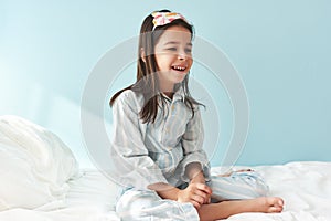Positive little girl wearing pajama in white stripes on blue, smiling broadly, sitting in the bed after wake up in the morning,