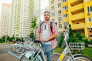 Positive man with a beard and bright casual clothes stands with a bicycle in the background of a row of bicycles and looks at the