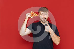 Positive man with beard stands on red background with piece of pizza in hand, looks into camera, smiles and shows thumb up. Hungry