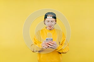 Positive man in a cap and casual clothes stands on a yellow background with a smile on his face and uses a smartphone