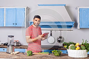 Positive man standing in modern kitchen holding tablet pc and looking at camera with smile, searching food recipe on internet,