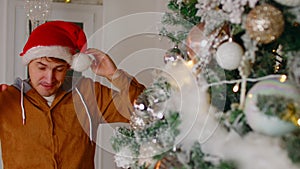 Positive man standing near Christmas tree. Young male puts santa hat. Happy man looking at camera and smiling while