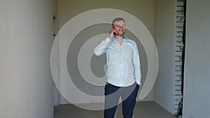 Positive man standing in unfurnished room in new flat picking up the phone talking and smiling. Portrait of confident