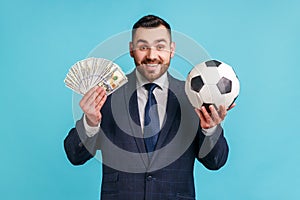 Positive man wearing official style suit holding soccer ball and hundred dollar bills, looking