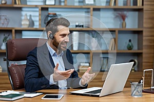 Positive man in wireless headset with micro hosting video conference while sitting by desktop with computer. Millennial