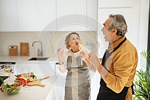 Positive married senior couple having fun while cooking in kitchen interior, singing and dancing, using untensils as mic