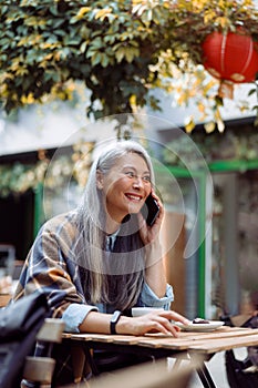 Positive mature Asian woman talks on cellphone at small table in outdoors cafe