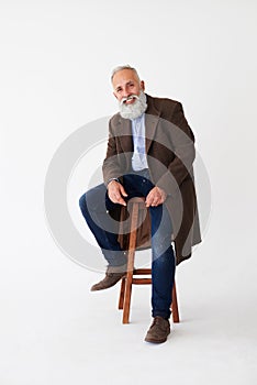 Positive mature bearded man in coat sitting on chair in studio
