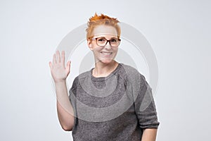 Positive mature caucasian woman with red hairstyle and glasses smiling friendly and waving hand at camera.