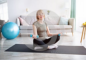 Positive mature woman in sports clothes doing strength exercises with dumbbells at home, free space