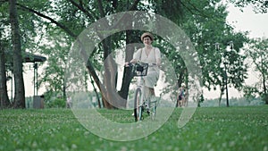 Positive mid-adult man and woman riding bikes on green lawn in summer park and leaving. Happy smiling elegant Caucasian