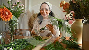 Positive middle age female florist preparing bouquet at table in flower shop. Floristry and small business concept
