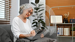 Positive middle age woman browsing wireless internet on digital tablet, relaxing on couch at home