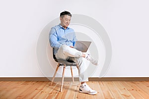 Positive middle aged asian man using laptop, sitting in chair against white studio wall, free space, full length