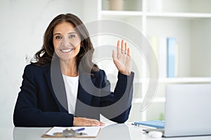 Positive middle aged businesswoman posing at office, waving at camera