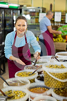 Positive middle-aged female merchandiser in apron picking up olives with scoop from plastic container in grocery store