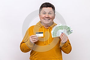 Positive middle aged man holding and showing euro cash and credit card, satisfied with cashback.