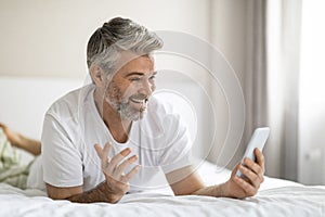 Positive middle aged man lying on bed, have video chat