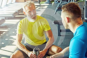 Positive middle aged man in sportswear holding bottle of water, talking with his personal trainer or fitness instructor