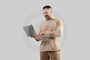 Positive middle aged man typing on laptop computer, surfing in internet, working online, standing on light background