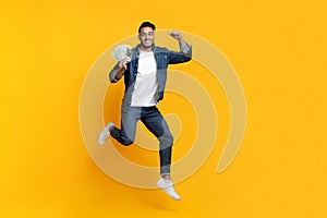 Positive middle-eastern man holding money on yellow background, celebrating win