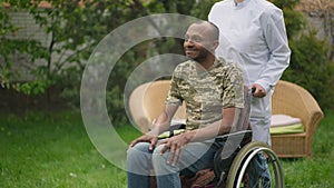 Positive military man smiling as unrecognizable nurse pushing wheelchair. Portrait of optimistic African American