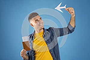 Positive millennial guy with paper plane, tickets and international passport on blue studio background