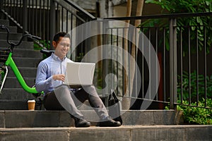 Positive millennial male entrepreneur sitting on the stairs in the city and using laptop