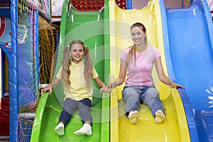 Positive millennial mother and her teenage daughter riding slide together at kids amusement centre