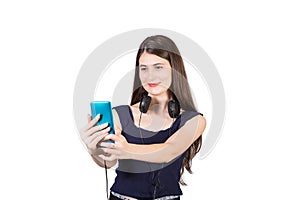 Positive millennial young woman with headset around her neck looks contented as takes a selfie with her phone, isolated on white