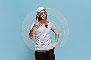Positive mood. Portrait of young smiling man with long hair posing in cap and sunglasses against blue studio background