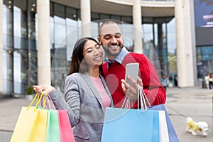Positive multiracial couple with bright gift bags and cellphone placing online order near shopping mall