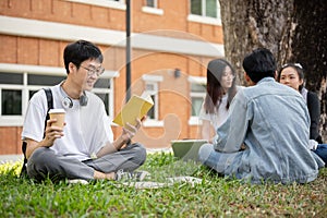 A positive and nerd Asian male college student is reading a book in a campus park with his friends