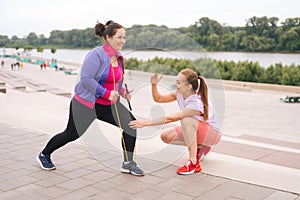 Positive obese overweight young woman doing exercises using fitness tape for weight loss with personal trainer outdoor