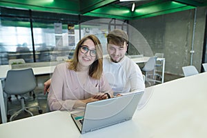 Positive office workers sit at the table near the laptop, look at the camera and smile