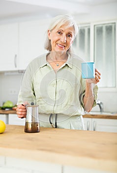 Positive old woman drinking coffee in kitchen at home