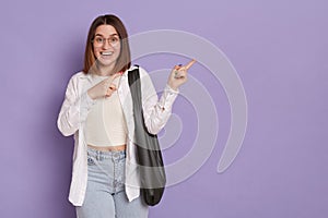 Positive optimistic woman with black bag wearing white shirt and jeans, standing and pointing aside showing copy space for