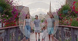 Positive parents and kids walking bridge with colorful flowers. Happy family spend time outdoors. Joyful couple with