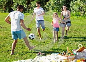 Positive parents with two kids playing soccer together on green field on summer day