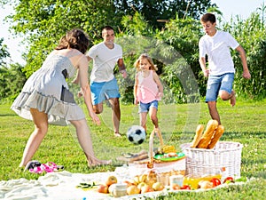 Positive parents with two kids playing soccer together on green field on summer day