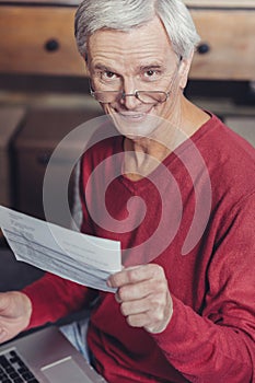 Positive pensioner smiling while holding a bill and a laptop