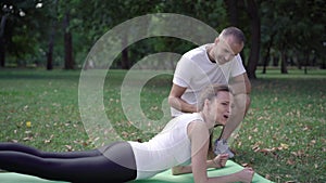 Positive personal coach controls sportswoman standing in plank position on exercise mat outdoors. Smiling handsome